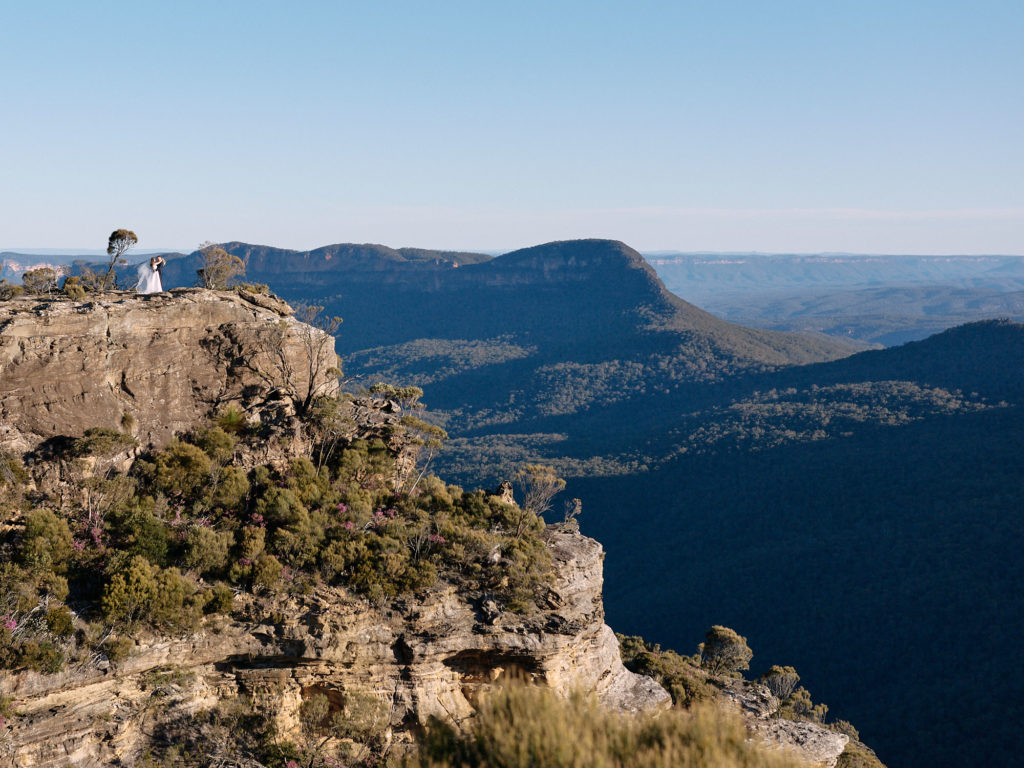 Wide shot of bride and groom standing on a rocky escarpment overlooking Mount Solitary and the Jamison Valley in the Blue Mountains