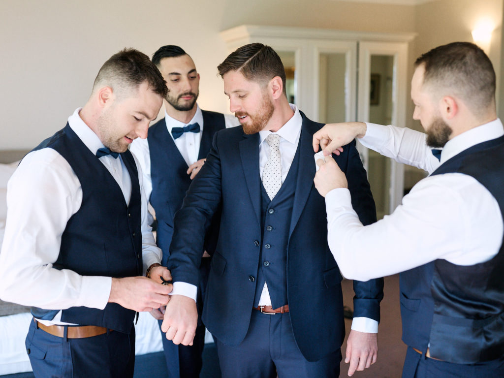 Groomsman helping to dress groom in his blue three piece suit on his wedding day in hotel room