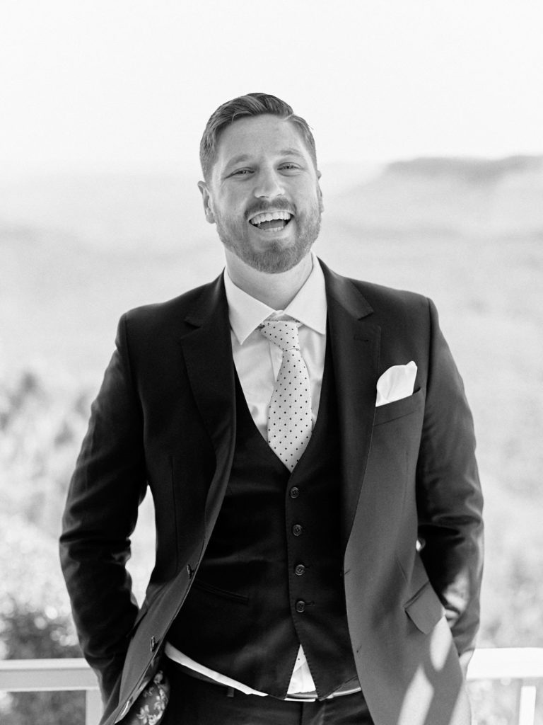 Black and white portrait of groom wearing three piece suit and tie, laughing on the balcony of his hotel room