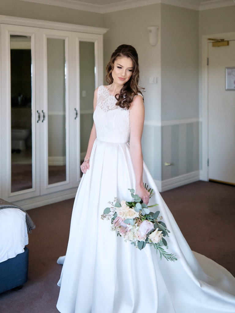 Full length photo of bride wearing her wedding dress and holding her bouquet, standing in front of wardrobe of hotel room