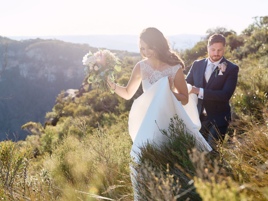 Groom holding brides dress off the ground while walking through long grass in the Blue Mountains